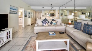 A living room with a sofa, coffee table, and a television. There is a kitchen in the background. - Navarre Beach Regency 705 - Nama' Stay At The Beach (Navarre)