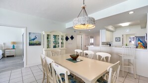 A dining area with a white table and chairs, a chandelier, and a cabinet with glass doors. - Navarre Beach Regency 705 - Nama' Stay At The Beach (Navarre)