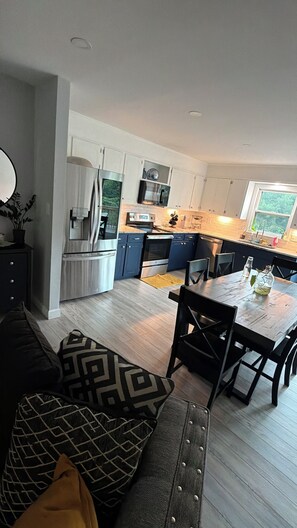A modern kitchen with white cabinets, stainless steel appliances, and a dining area with a wooden table and chairs. - Relax at Hibiscus Grove - w/ an Amazing Deck View (Kings Mountain)