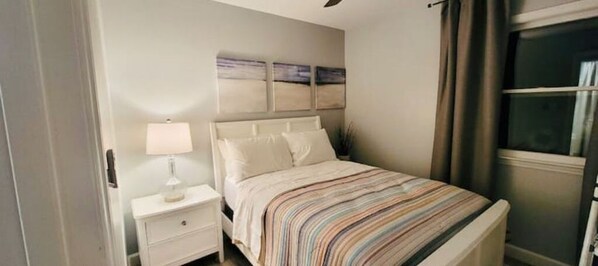 A bedroom with a bed, nightstand, lamp, and artwork on the wall. - Relax at Hibiscus Grove - w/ an Amazing Deck View (Kings Mountain)