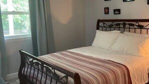 Et soveværelse med seng, ventilator, et vindue med gardiner og billeder i rammer på væggen.