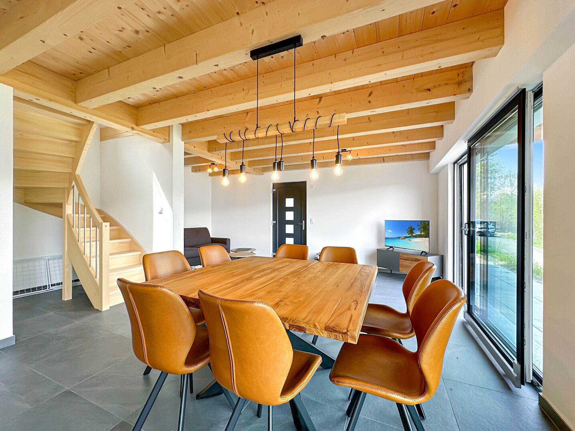 A modern dining area with a wooden table and leather chairs, a staircase, and a large window with a view.