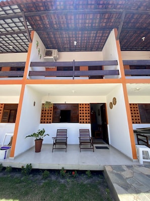 A two-story building with a covered patio area, two wooden chairs, a potted plant, and a small garden. - Casa duplex na Praia do Francês (Marechal Deodoro)