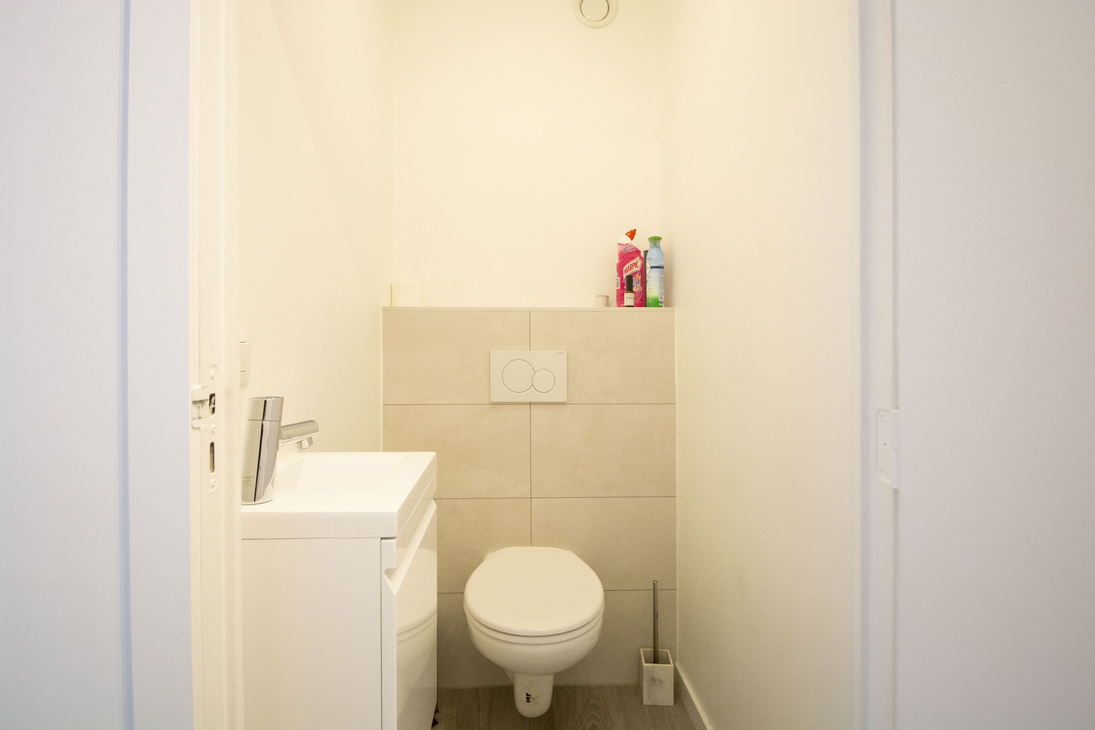 Un baño pequeño y limpio, con inodoro, lavabo y repisa con artículos de aseo.