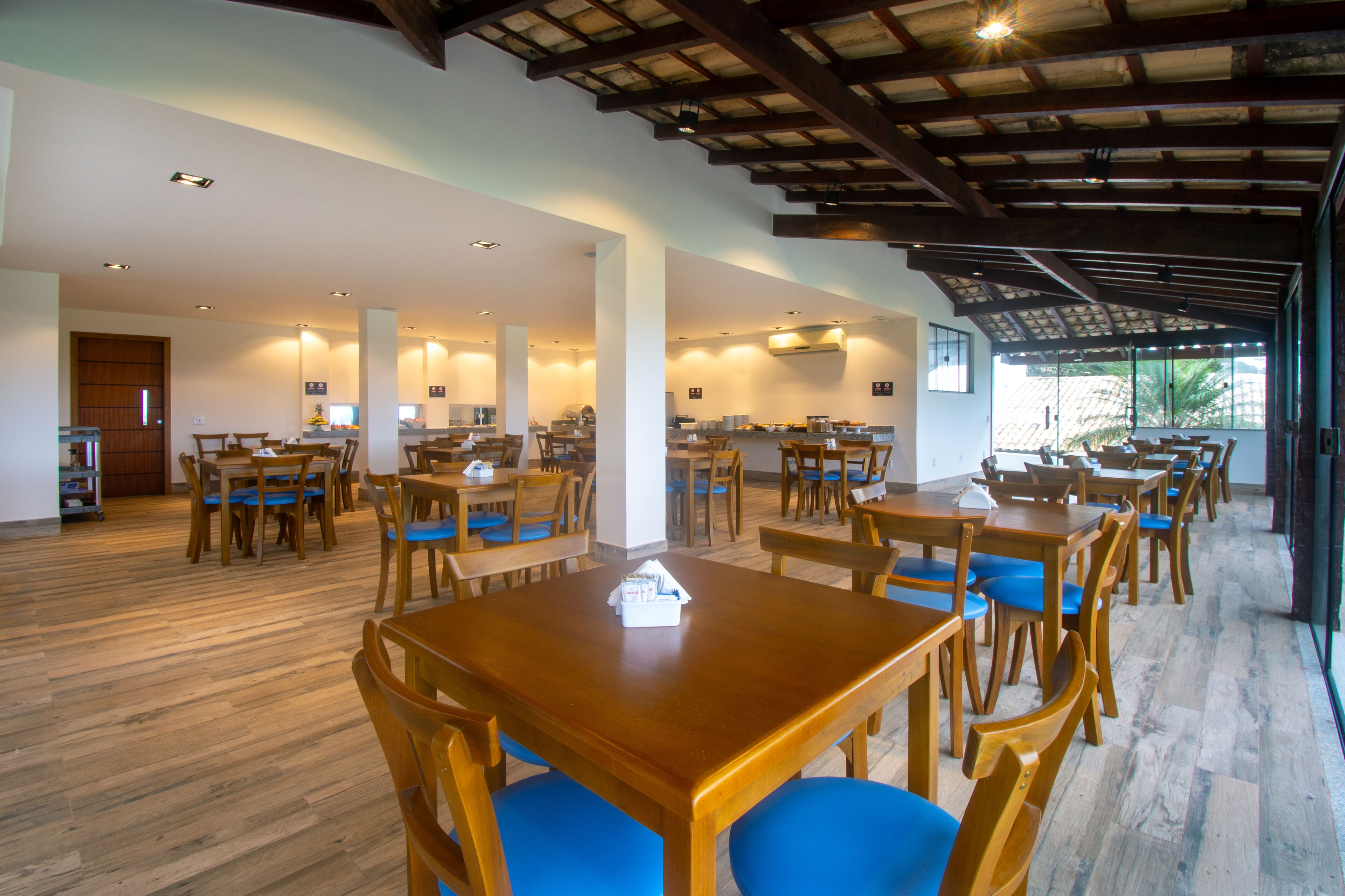 Un comedor espacioso con mesas de madera y sillas azules, que cuenta con un techo alto con vigas a la vista y luz natural gracias a grandes ventanales.