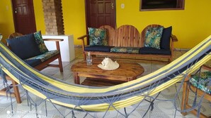 A room with a yellow wall, a wooden bench with cushions, a hammock, and a small table with a bottle and a hat. - POUSADA ÁGUA DE COCO (Prado)
