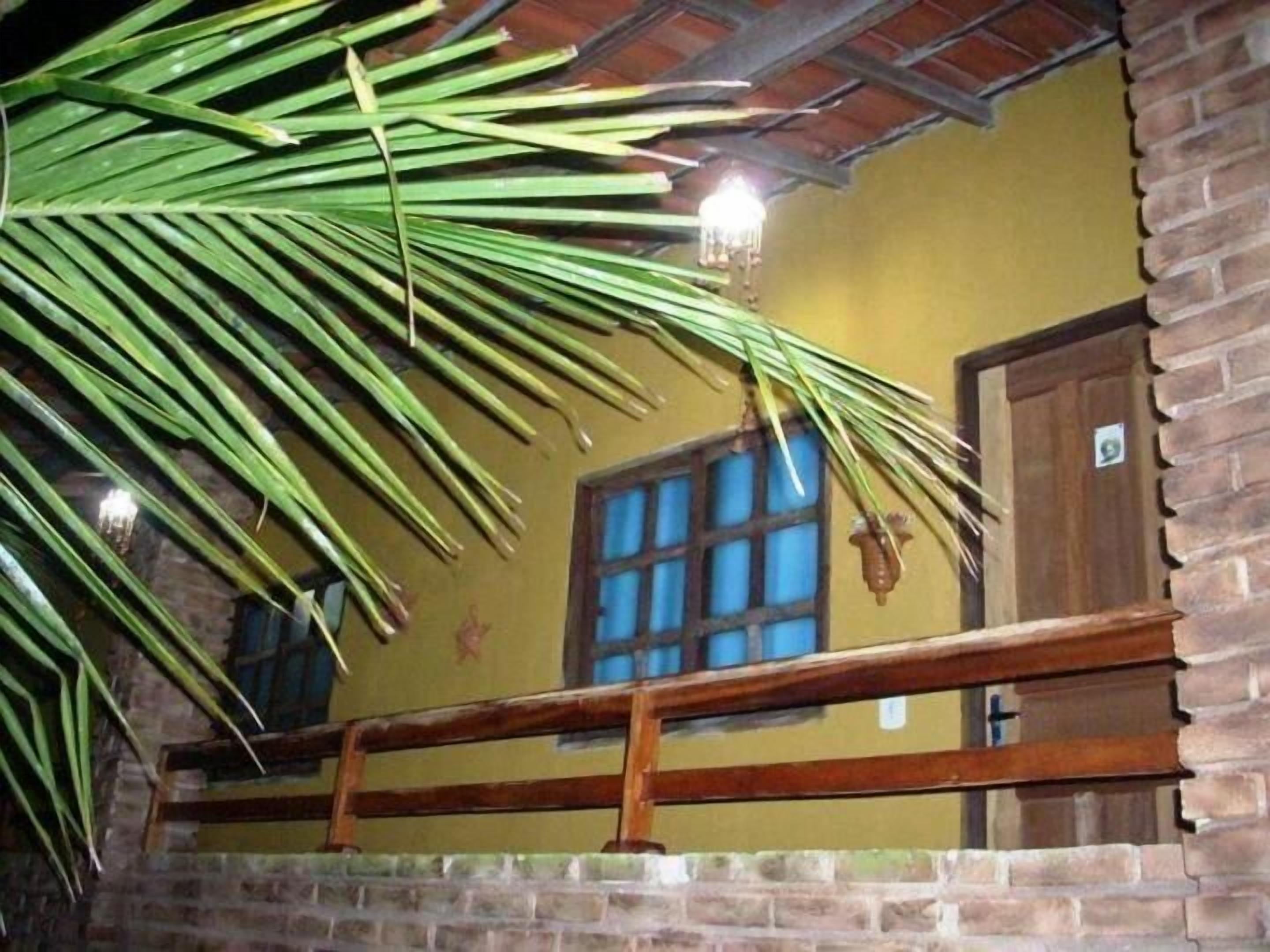 A room with a brick wall, a wooden railing, a window with blue panes, and a palm leaf hanging from the ceiling.