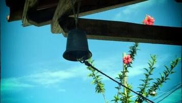 A bell hanging from a wooden structure with a clear blue sky and a plant with pink flowers in the background.