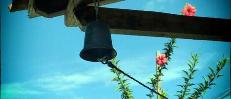 A bell hanging from a wooden structure with a clear blue sky and a plant with pink flowers in the background.