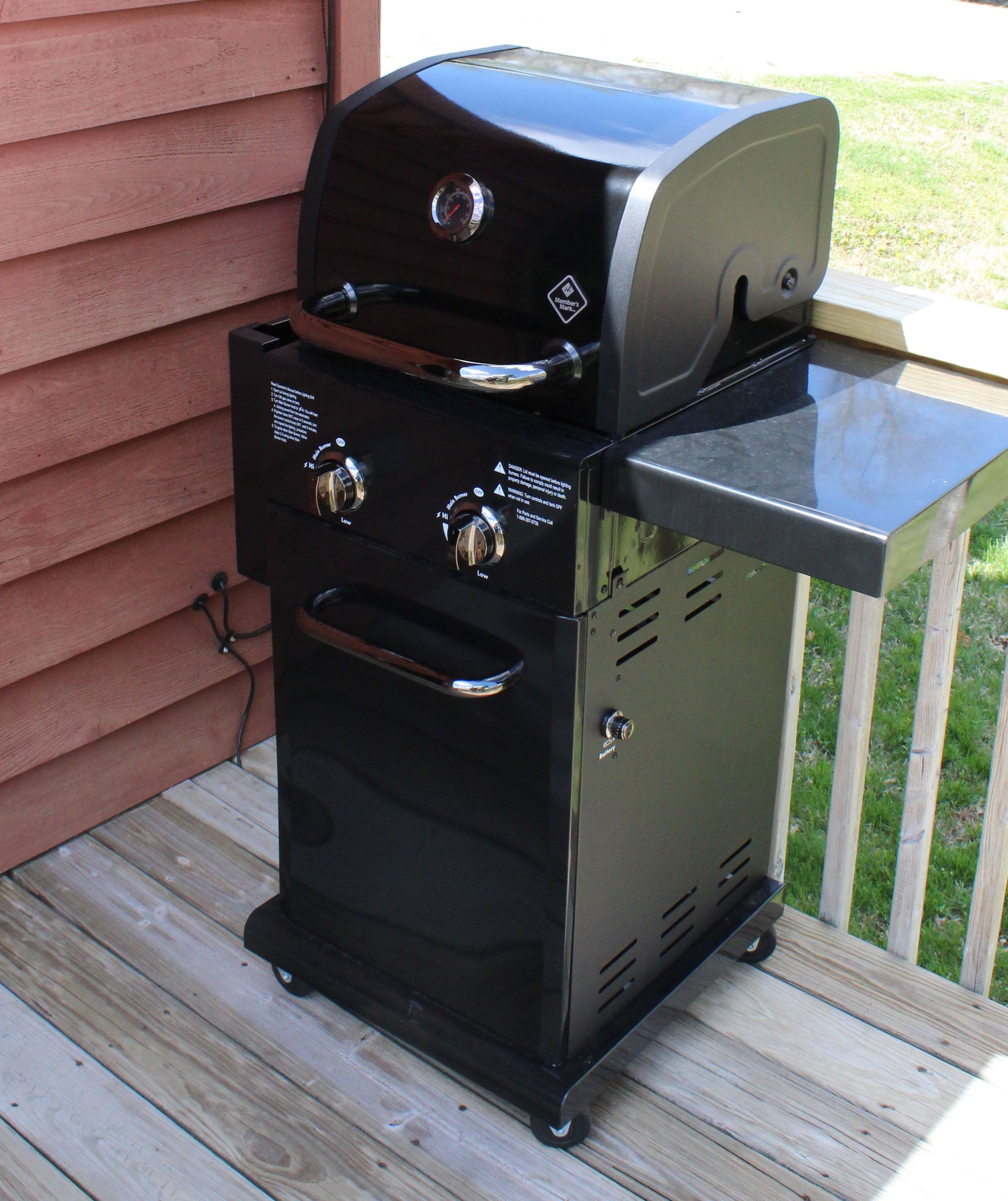 Una parrilla negra para barbacoa con dos perillas de control y un quemador lateral, ubicada sobre una terraza de madera.