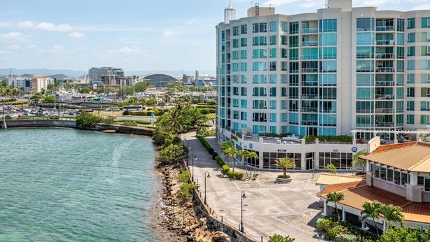 A modern hotel with a curved facade, located near a waterfront with a paved walkway and greenery. - Panoramic Lagoon Studio · Water Backup (San Juan)