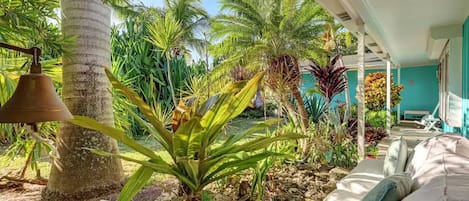 A tropical garden with a stone pathway, a bell, and a white sofa with cushions.