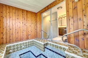 A wooden interior with a small hot tub area, a shower, and a bathroom with a mirror and sink.