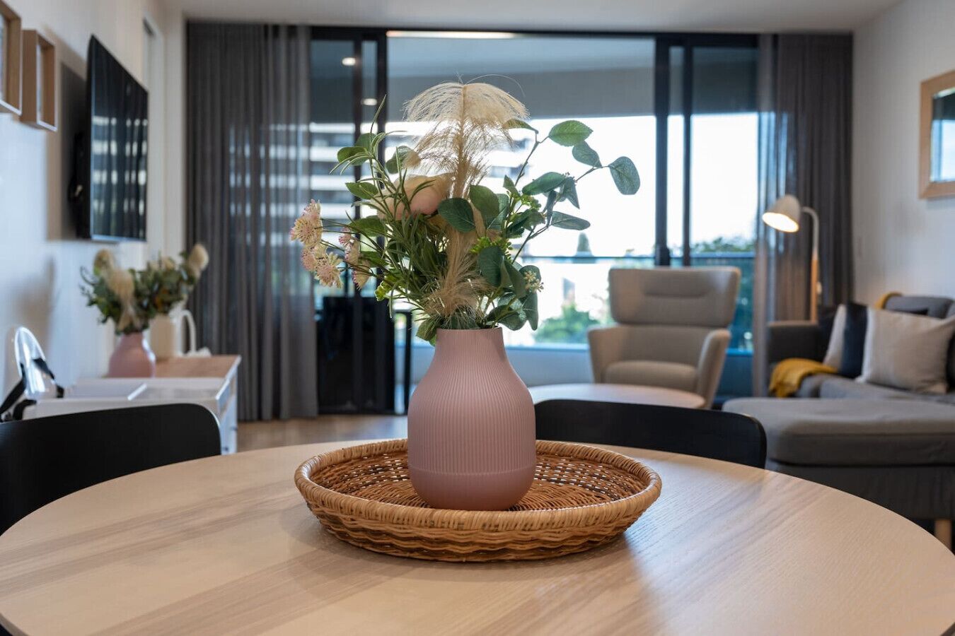 Una sala de estar moderna con una mesa redonda, un florero rosa con flores y vistas al exterior a través de amplios ventanales.