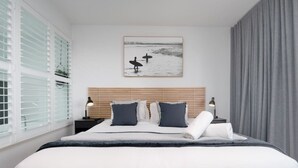 A modern hotel room with a large bed, wooden headboard, and a framed picture of surfers on the beach.