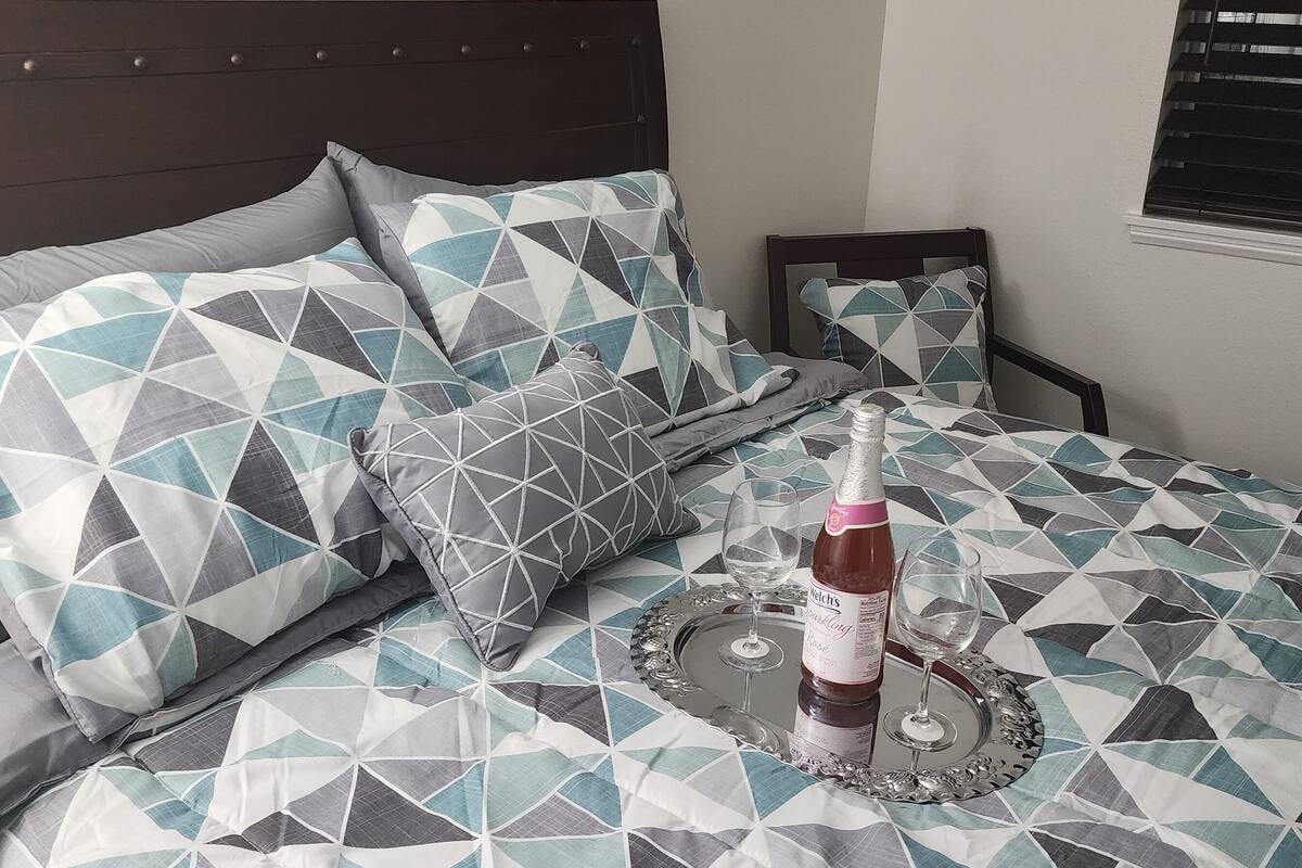 Un lit bien fait, avec une literie à motifs géométriques, un plateau avec deux verres et une bouteille de vin, et une fenêtre avec des stores.