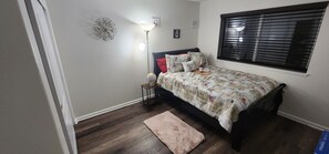 A bedroom with a bed, a nightstand, a lamp, a window with blinds, and a decorative wall piece. - CSE Property Group (Simi Valley)