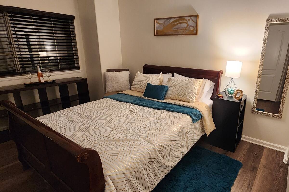 Une chambre à coucher avec un lit, une table de chevet, une lampe, un miroir et une fenêtre avec des stores.