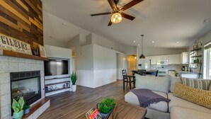 A spacious living area with a fireplace, a ceiling fan, and a kitchen in the background.