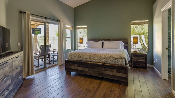 A bedroom with a large bed, wooden flooring, a TV, and a sliding glass door leading to an outdoor area.