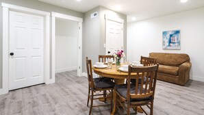 A dining area with a wooden table set for four, surrounded by chairs with cushions. There is a brown sofa against the wall and a white door to the left.