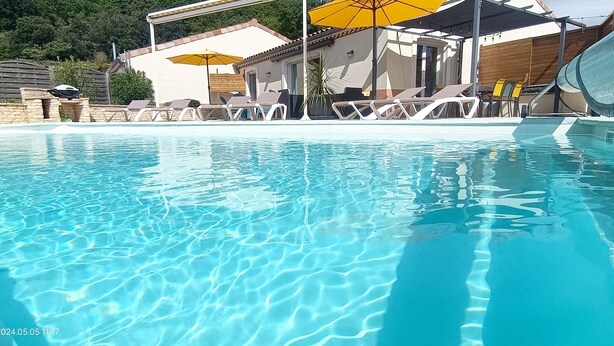 Una piscina cristalina con sillas de descanso y sombrillas, rodeada por una valla de madera y un edificio con techo de tejas.