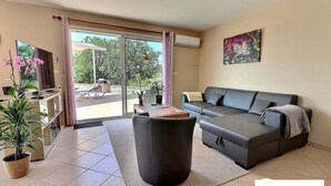 Una sala de estar moderna con un sofá, una mesa de centro y un televisor. Posee un ventanal corredero que da a un espacio exterior con una barbacoa.
