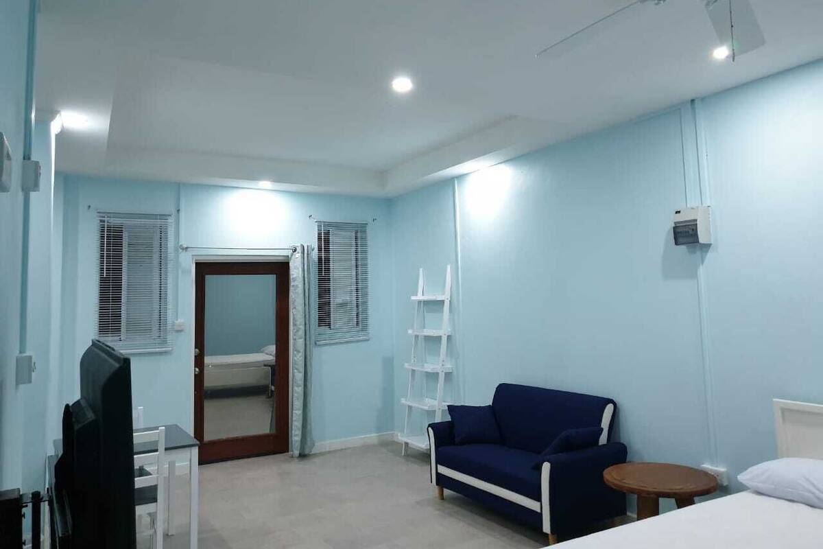 A room with a blue wall, a white ceiling fan, a white shelf, a blue sofa, a white bedside table, and a door with a mirror.