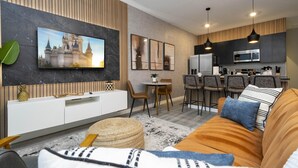 Una sala moderna con un televisor de pantalla plana grande, un sofá, un comedor con sillas y una cocina con microondas y refrigerador.