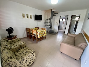 A living room with a sofa, a dining table with chairs, a TV mounted on the wall, and a staircase leading to another level.