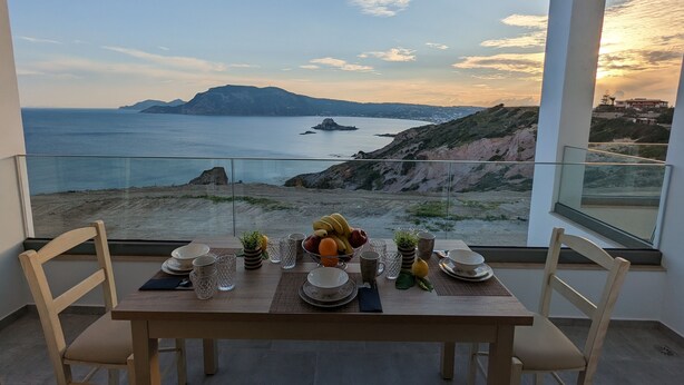 A dining table set with plates, glasses, and fruits, overlooking a coastal landscape with cliffs and the sea. - Cute Apartments (Κέφαλος)