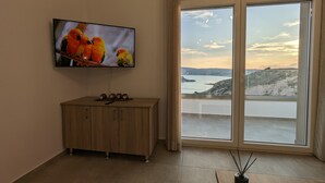A room with a TV displaying colorful parrots, a wooden cabinet, and a large window offering a coastal view. - Cute Apartments (Κέφαλος)