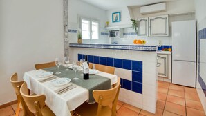 A dining area with a table set for two, a kitchen with blue and white tiles, and a white refrigerator. - LES HUPPES: Nice ground floor house in l'Escala with air-conditioning and large terrace (L'Escala)