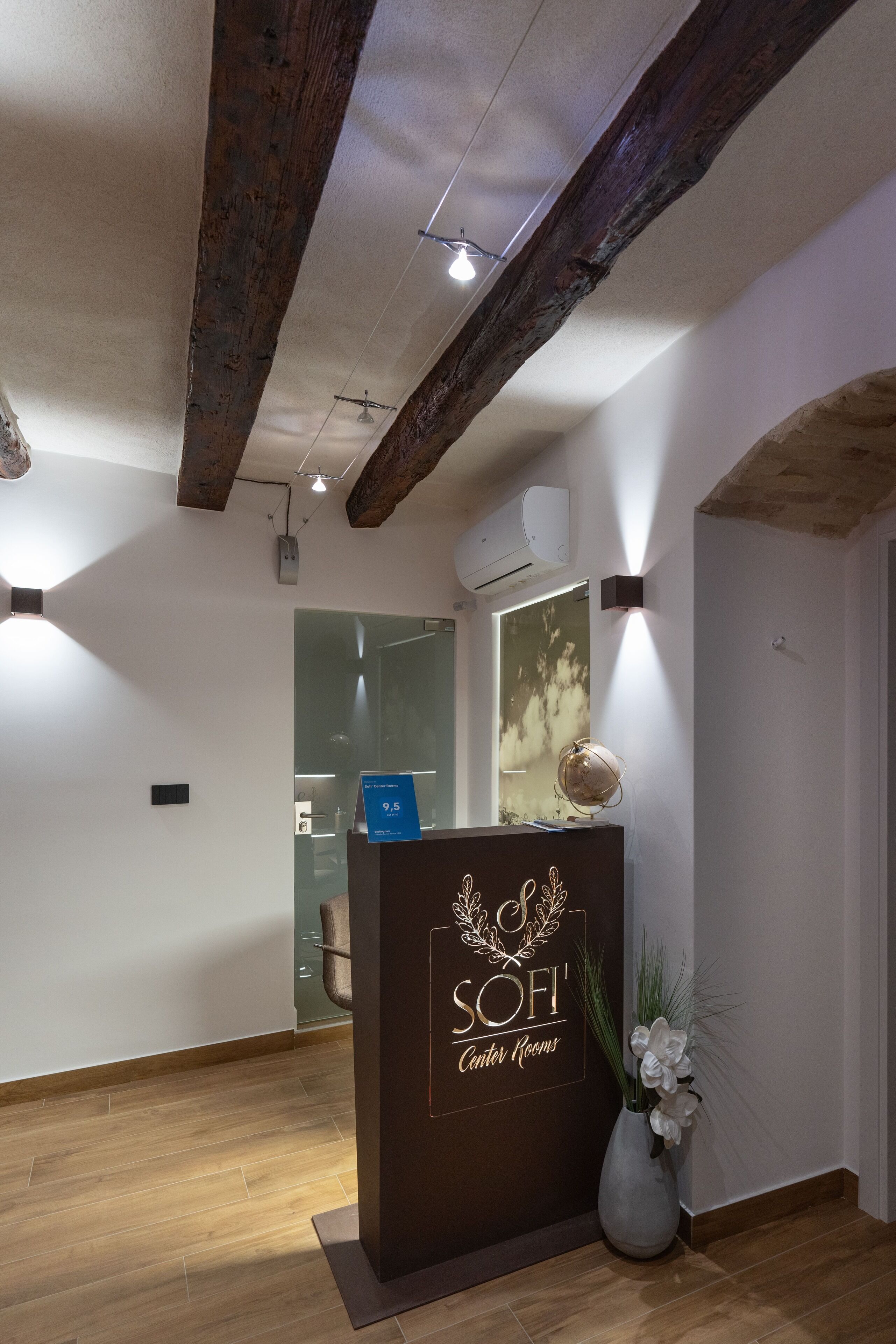 Una zona de recepción con techo de madera, un mostrador de recepción que dice "SOFI Center Rooms" y un jarrón con flores blancas.