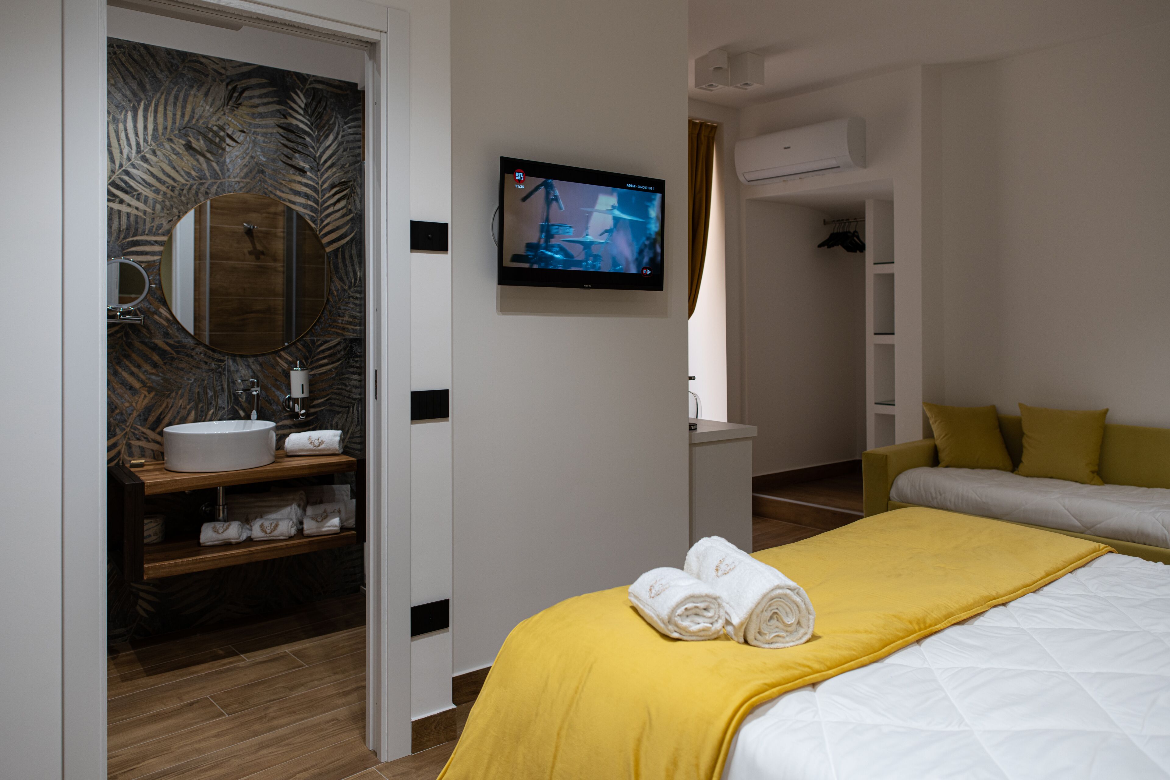 Una habitación de hotel moderna con un tocador de madera, un espejo grande, un televisor de pantalla plana, una cama con una manta amarilla y un sofá.
