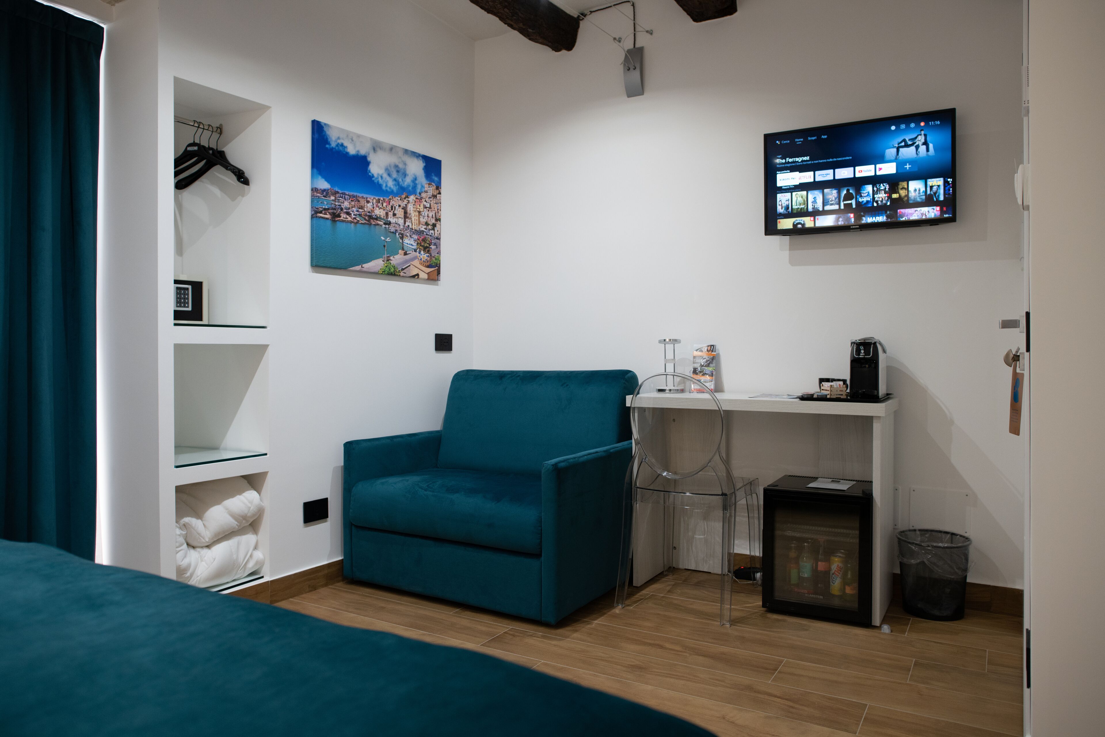A modern hotel room with a teal sofa, a flat-screen TV mounted on the wall, a small desk with a coffee machine, and a view of a coastal cityscape.