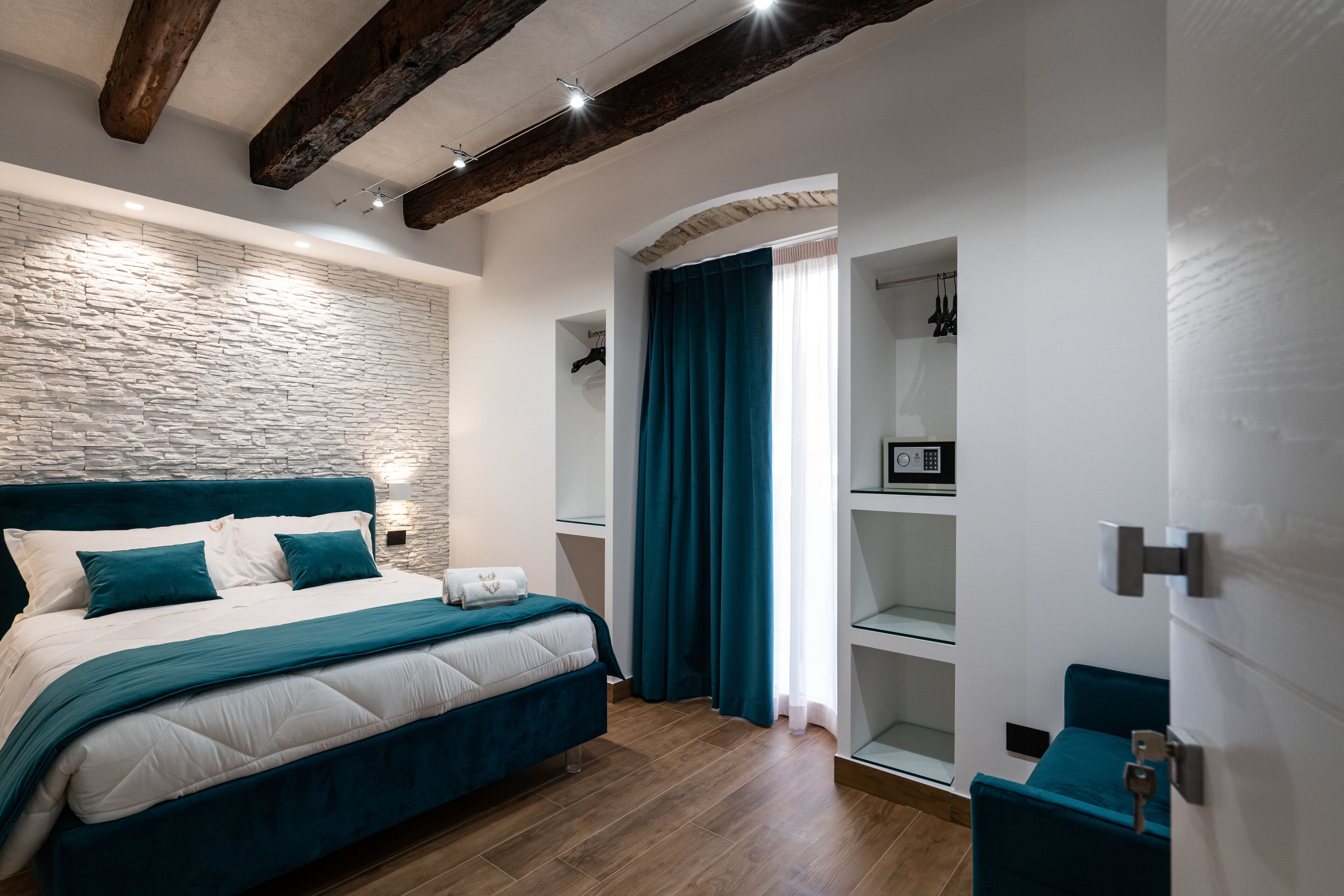 Une chambre avec un mur en pierre, des poutres apparentes, un lit avec une literie de couleur turquoise, une armoire et un petit coin salon.