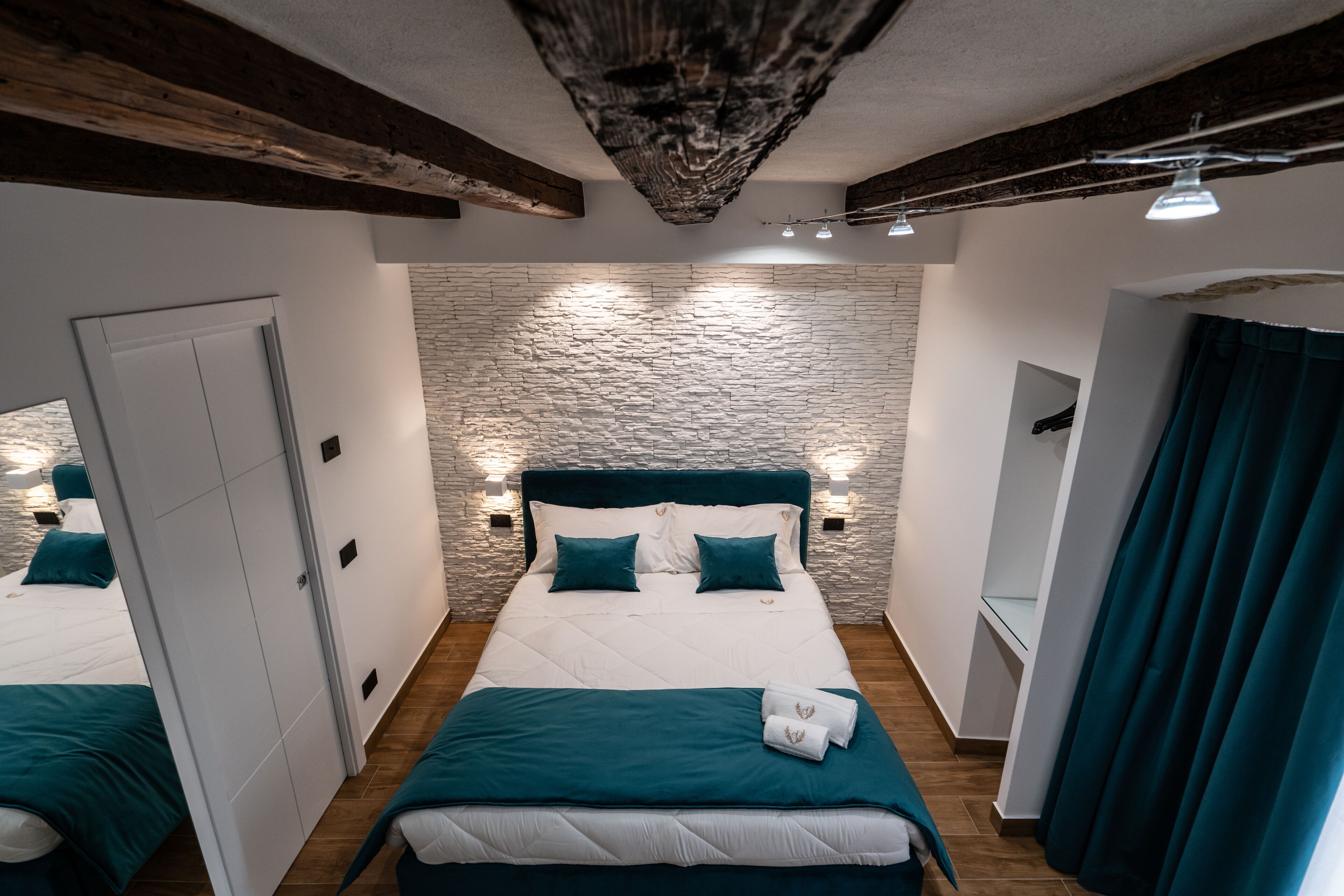 A bedroom with a stone wall, a wooden ceiling, a bed with teal bedding, and a mirror with curtains.