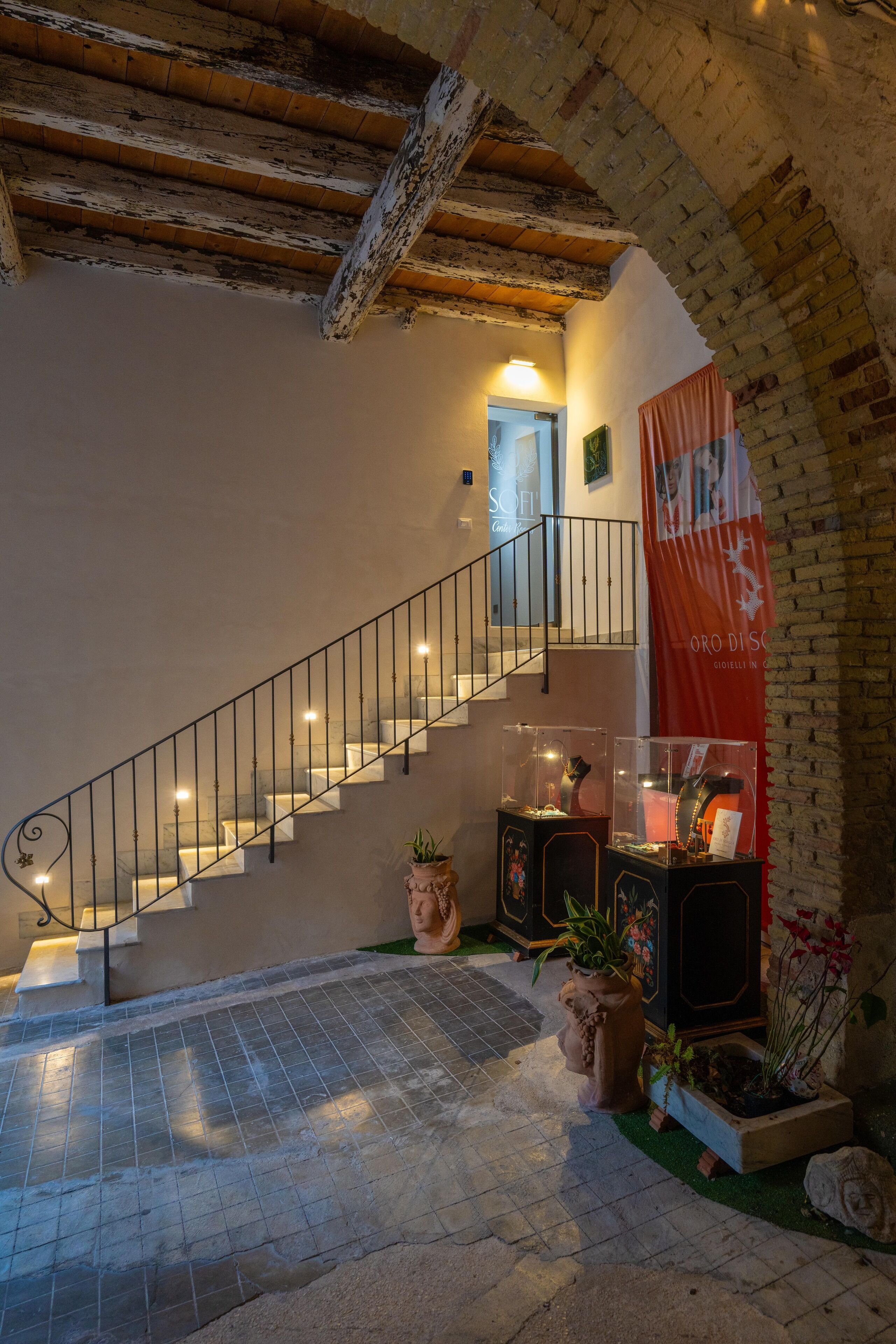 Un escalier avec une rampe en métal mène à un niveau supérieur. Au niveau inférieur, on trouve des plantes en pot et une vitrine exposant divers objets.