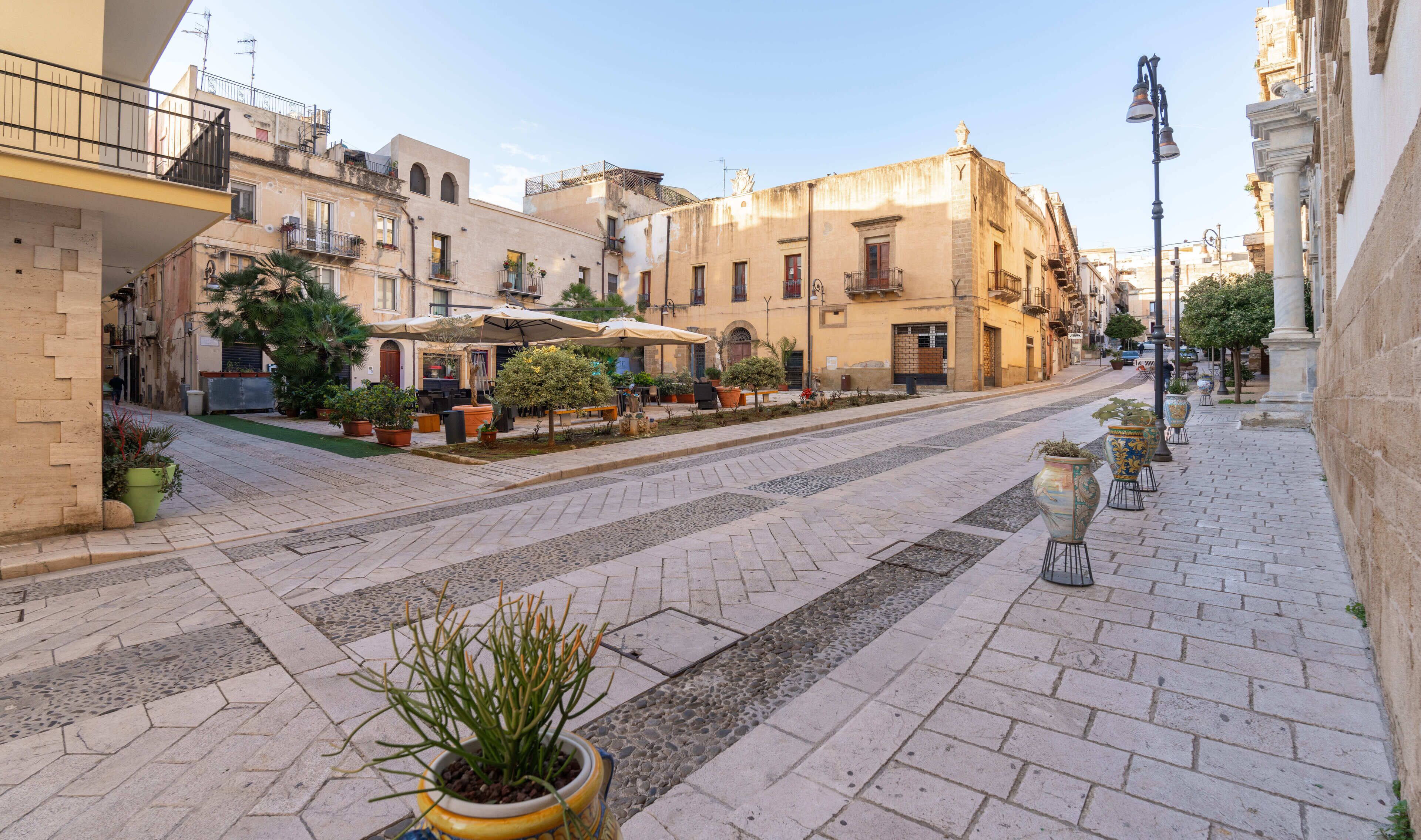 Una calle flanqueada por edificios, con mesas al aire libre y macetas.