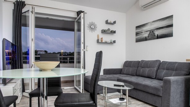A modern living room with a glass dining table, black chairs, a grey sofa, and a view through a sliding glass door. - Apartment Mia - Two Bedroom Apartment with Balcony (Split)
