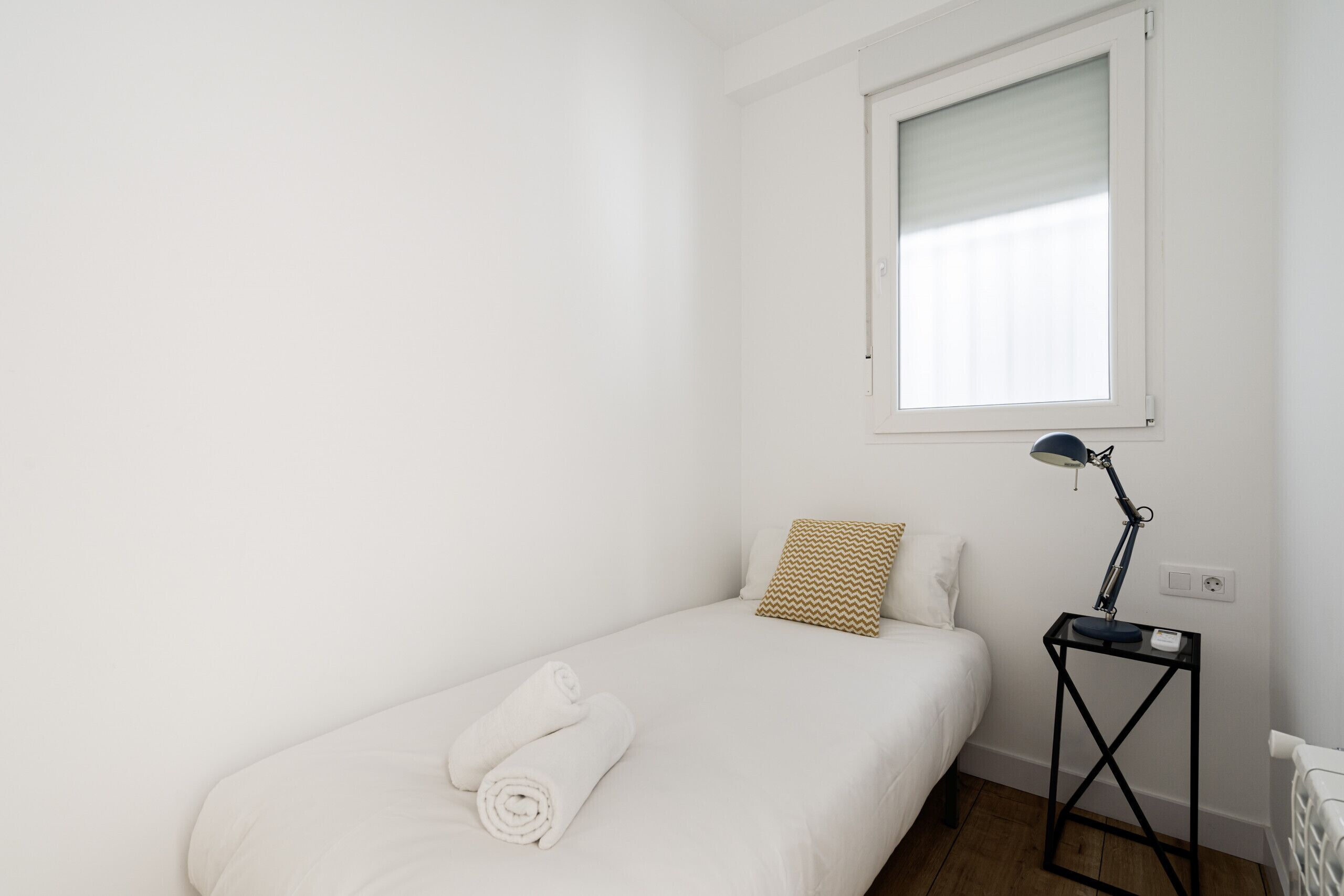 Een kleine, witte kamer met een enkel bed, een raam met zonwering, een klein tafeltje met een lamp en een radiator.