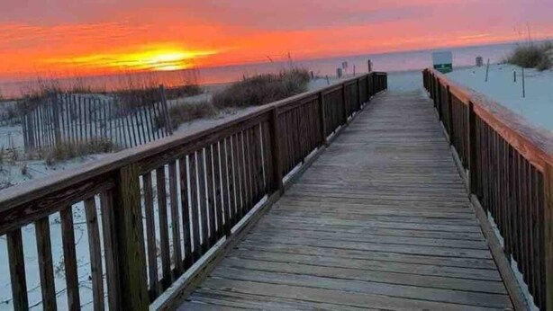 Une promenade en bois menant à une plage, avec un coucher de soleil en arrière-plan.