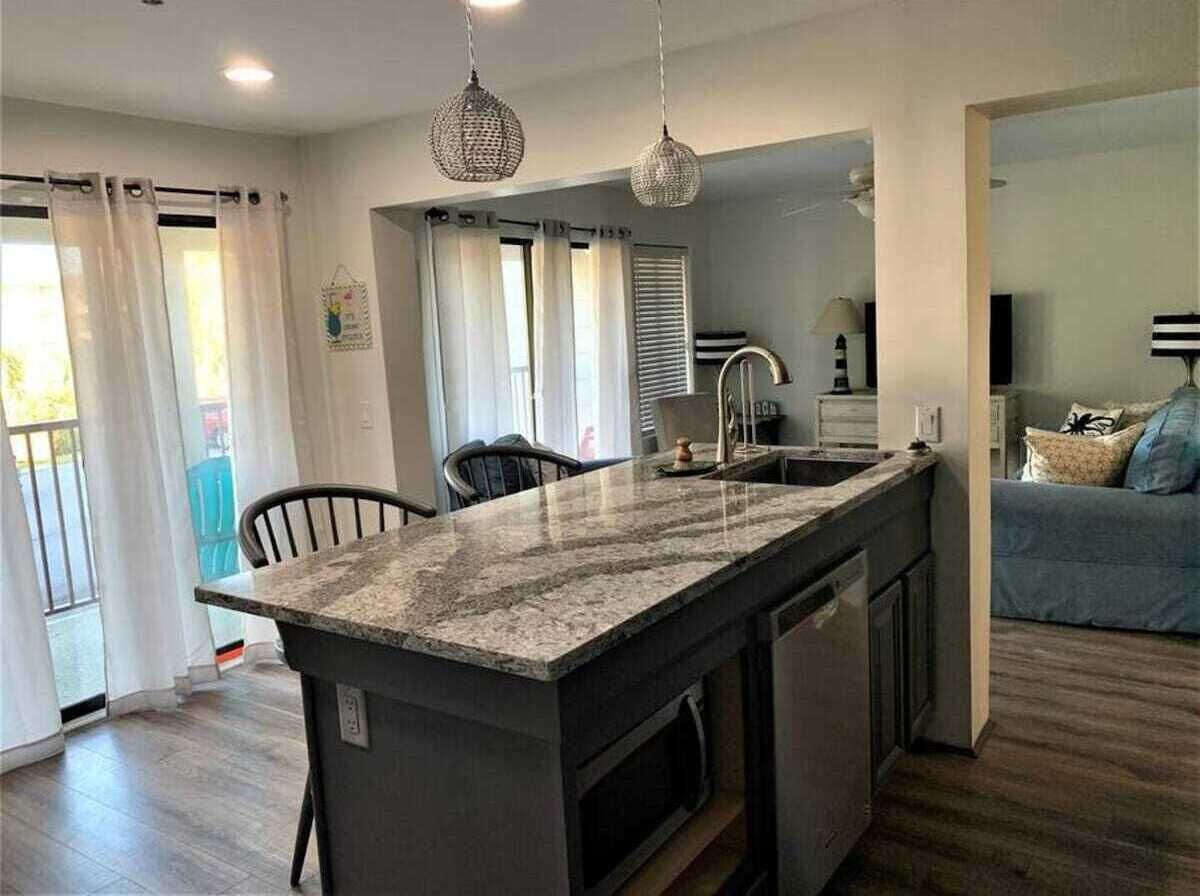 Una cucina moderna con piano di lavoro in granito, lavastoviglie integrata e lavello. C'è un'area pranzo con una sedia e un tavolo, e un soggiorno con divano e televisore.