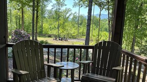 A wooden deck with two Adirondack chairs and a small table, overlooking a forested area with a picnic table in the distance. - Beautiful Mountain View with Large Saltwater Pool (Landrum)