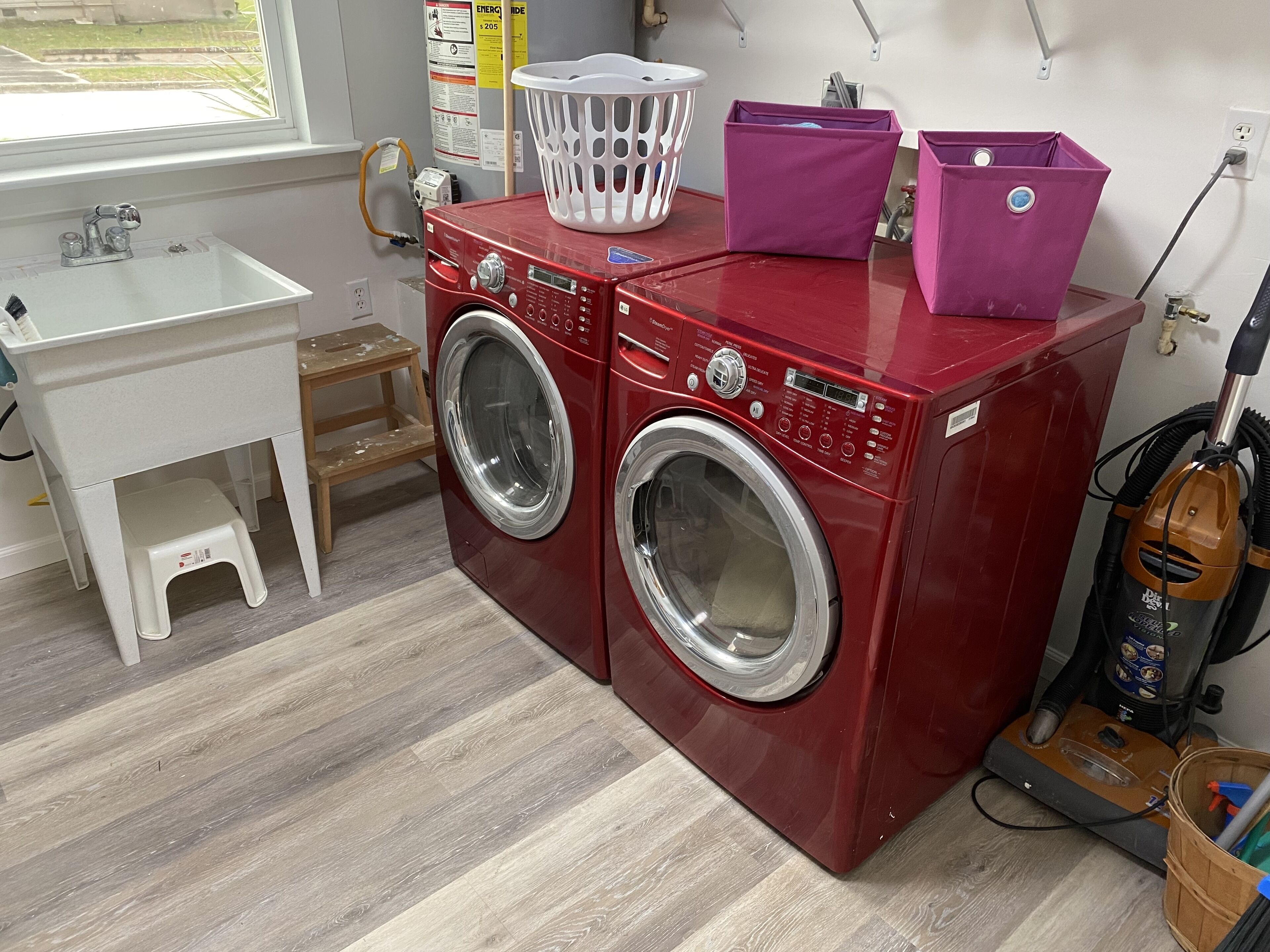 A laundry area with two red front-loading washing machines, a white sink, a wooden stool, a white laundry basket, and a vacuum cleaner.