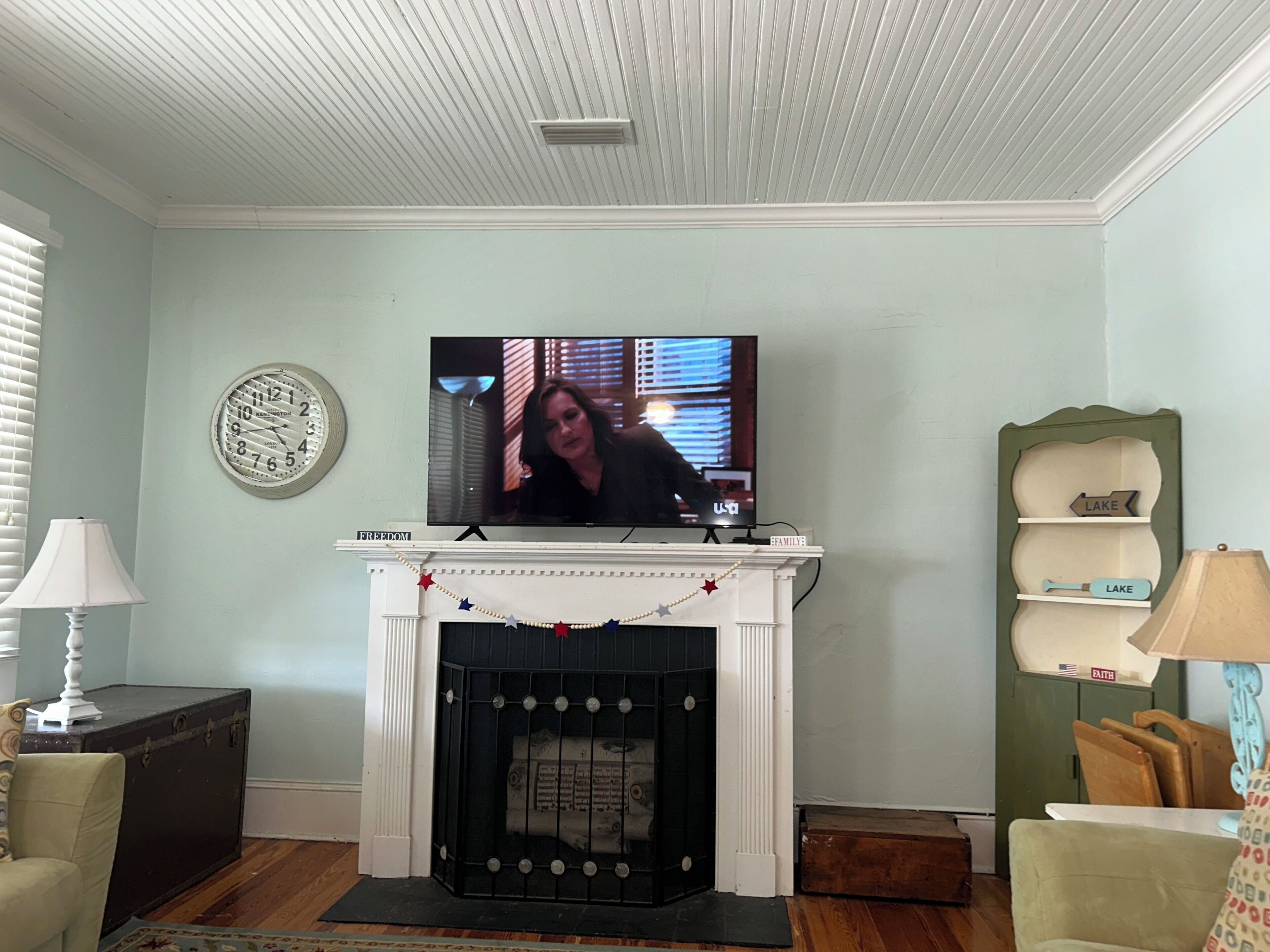 A living room with a fireplace, a television mounted above it, a clock on the wall, and a small shelf with decorative items.