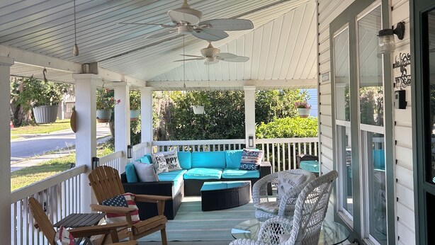 A covered porch with ceiling fans, a blue sofa with cushions, rocking chairs, and a glass table.