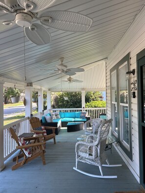 A covered porch with ceiling fans, a blue sofa with cushions, rocking chairs, and a glass table.