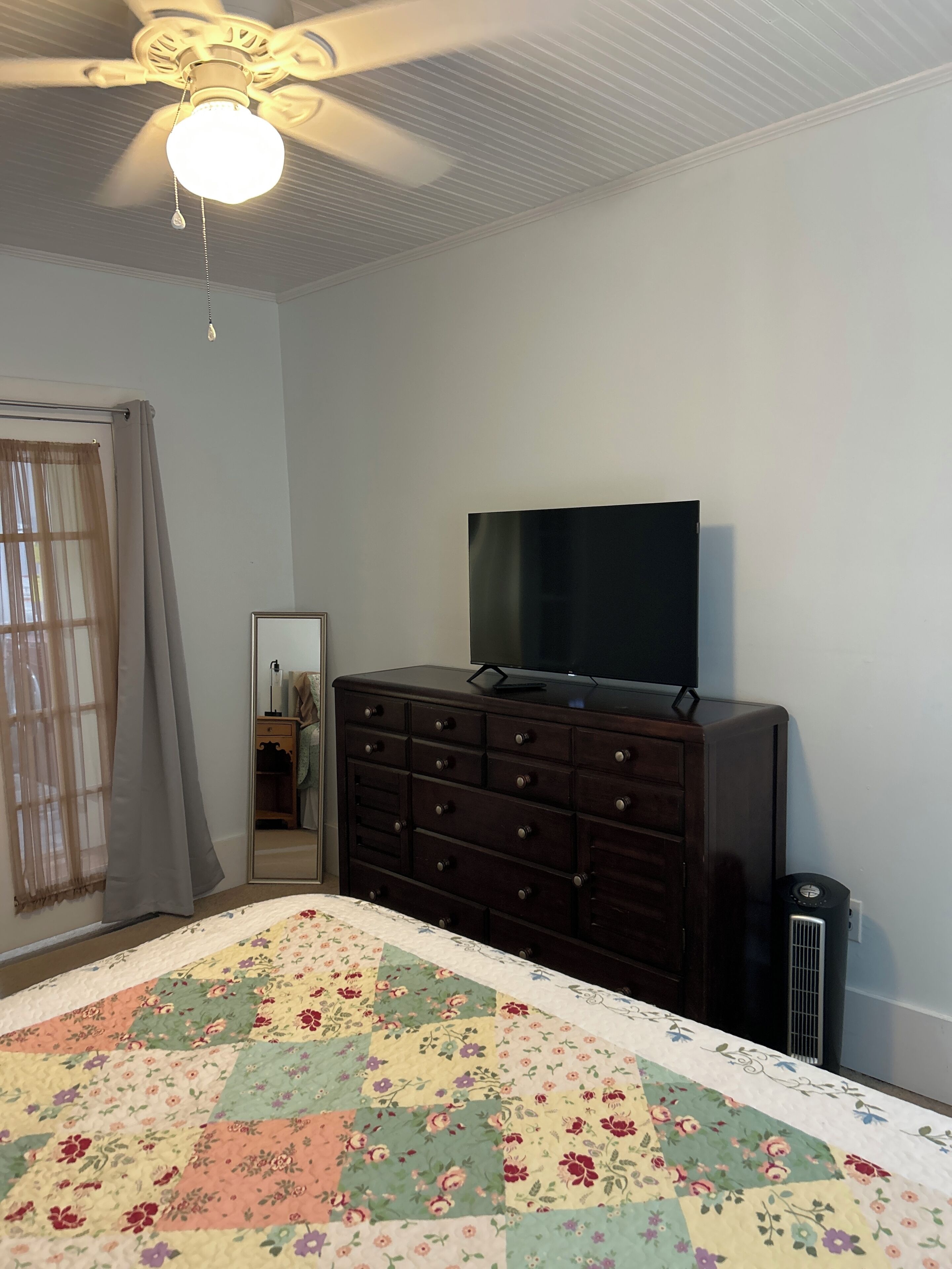 Una camera da letto con un letto, una cassettiera, un televisore sopra e un ventilatore a soffitto, oltre a una finestra con tende.